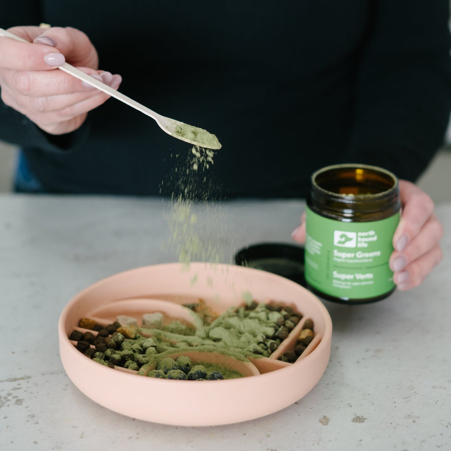 Person adding super green supplement for dogs from a spoon into a pink bowl with a green jar labeled 'Super Greens' on a grey surface.