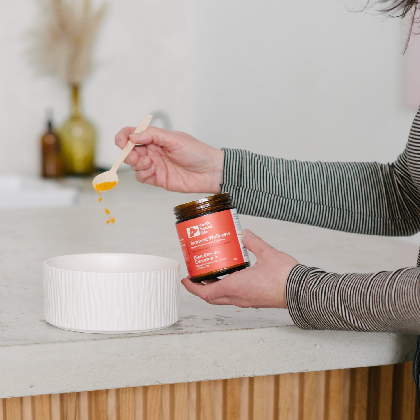 Person holding a jar of Turmeric Wellness+ supplement for dogs with a dog looking on in a kitchen setting