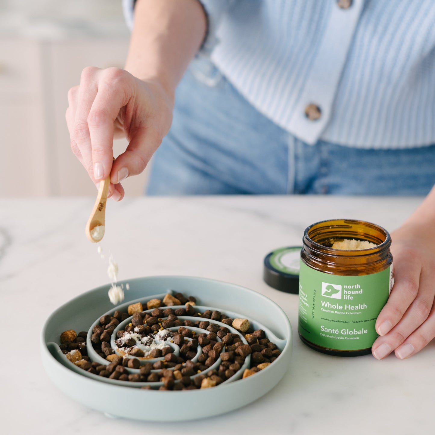 Person adding whole health supplement from a jar into a bowl of dog food on a table.