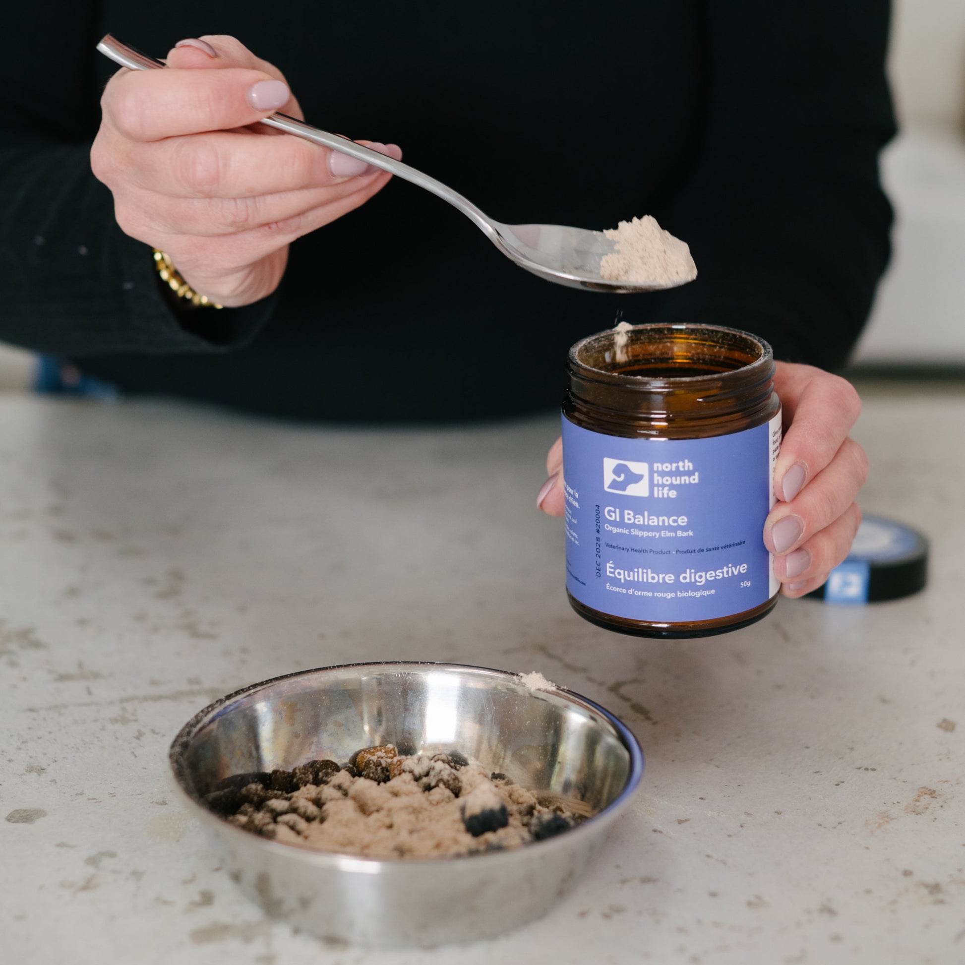 Person using a spoon to scoop a slippery elm bark powder from a jar labeled 'GI Balance' on a countertop.