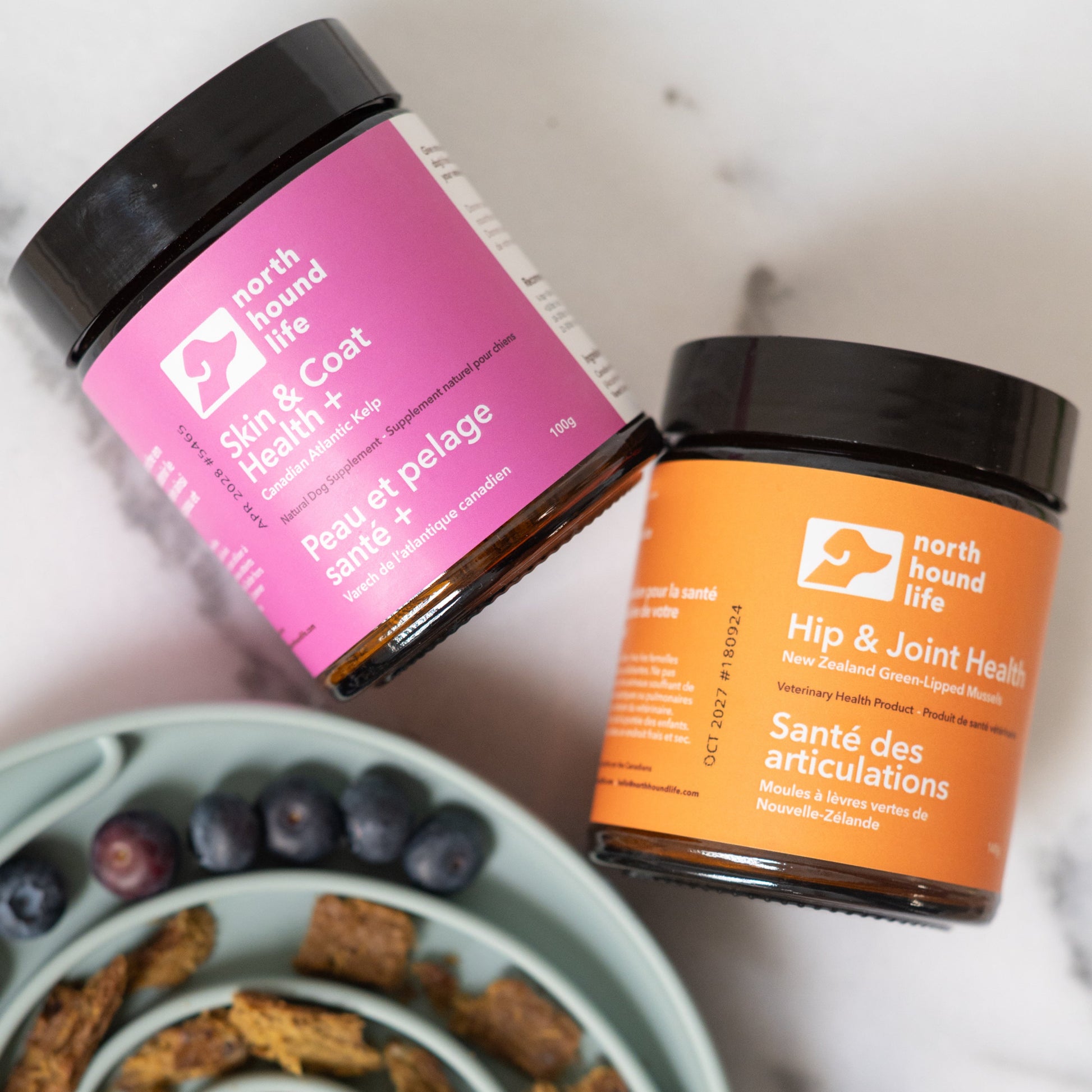 Two pet health supplement jars on a marble surface with a bowl of treats.